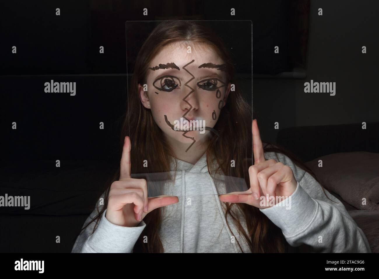 La teenager tiene un foglio di perspex trangenitore con il disegno di mezzo volto sul proprio volto per mostrare emozioni e sentimenti diversi Foto Stock
