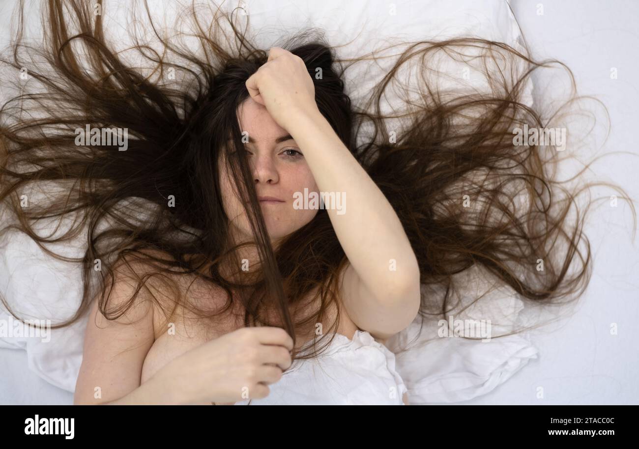 vista dall'alto di una donna bruna attraente, giovane, sexy, capelli selvaggi sulle lenzuola, con uno sguardo diretto, giocando con i suoi capelli, rilassata, distesa fresca e morbida Foto Stock