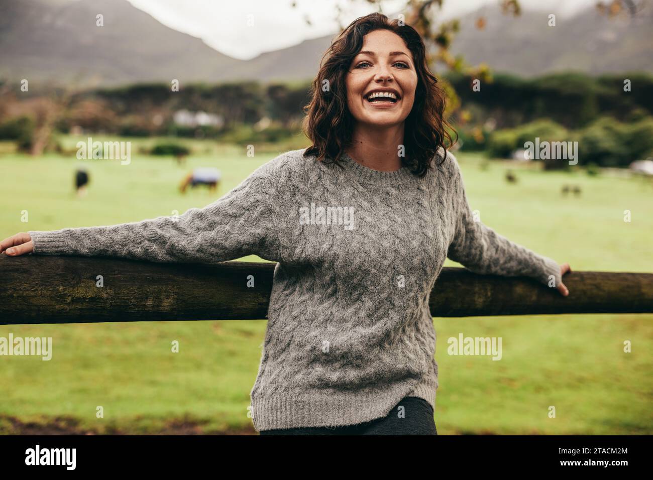Ritratto di donna allegra appoggiandosi a un recinto in campagna. La donna che indossa un maglione in piedi da una recinzione guardando la fotocamera e ridere. Foto Stock