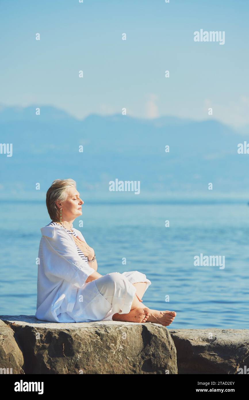 Ritratto all'aperto di una donna matura felice che medita vicino al lago o al mare, indossa abiti bianchi, spiritualità, risveglio ed equilibrio di vita Foto Stock