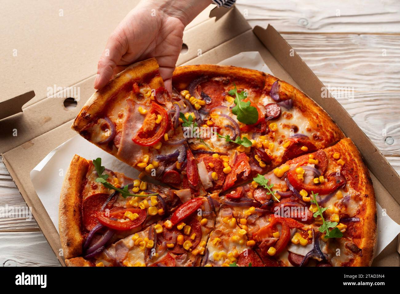 La mano caucasica umana prende una fetta di pizza grande dalla scatola di cartone su un tavolo di legno bianco Foto Stock