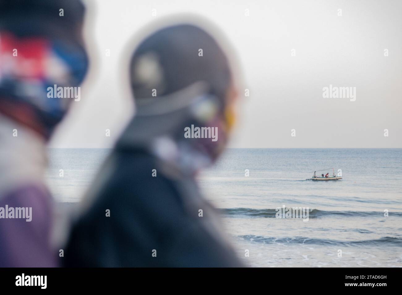 Due figure sfocate guardano al mare mentre una barca da pesca lontana naviga su acque calme sotto un cielo morbido e coperto Foto Stock