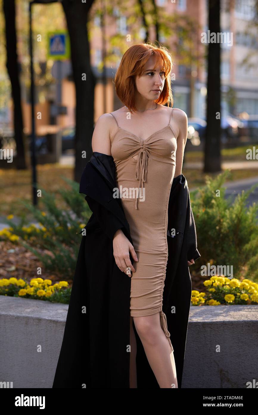 Una donna alla moda rossa che si gode una serata autunnale in città. Indossa un cappotto nero alla moda e un abito beige, per un'audace dichiarazione di stile Foto Stock