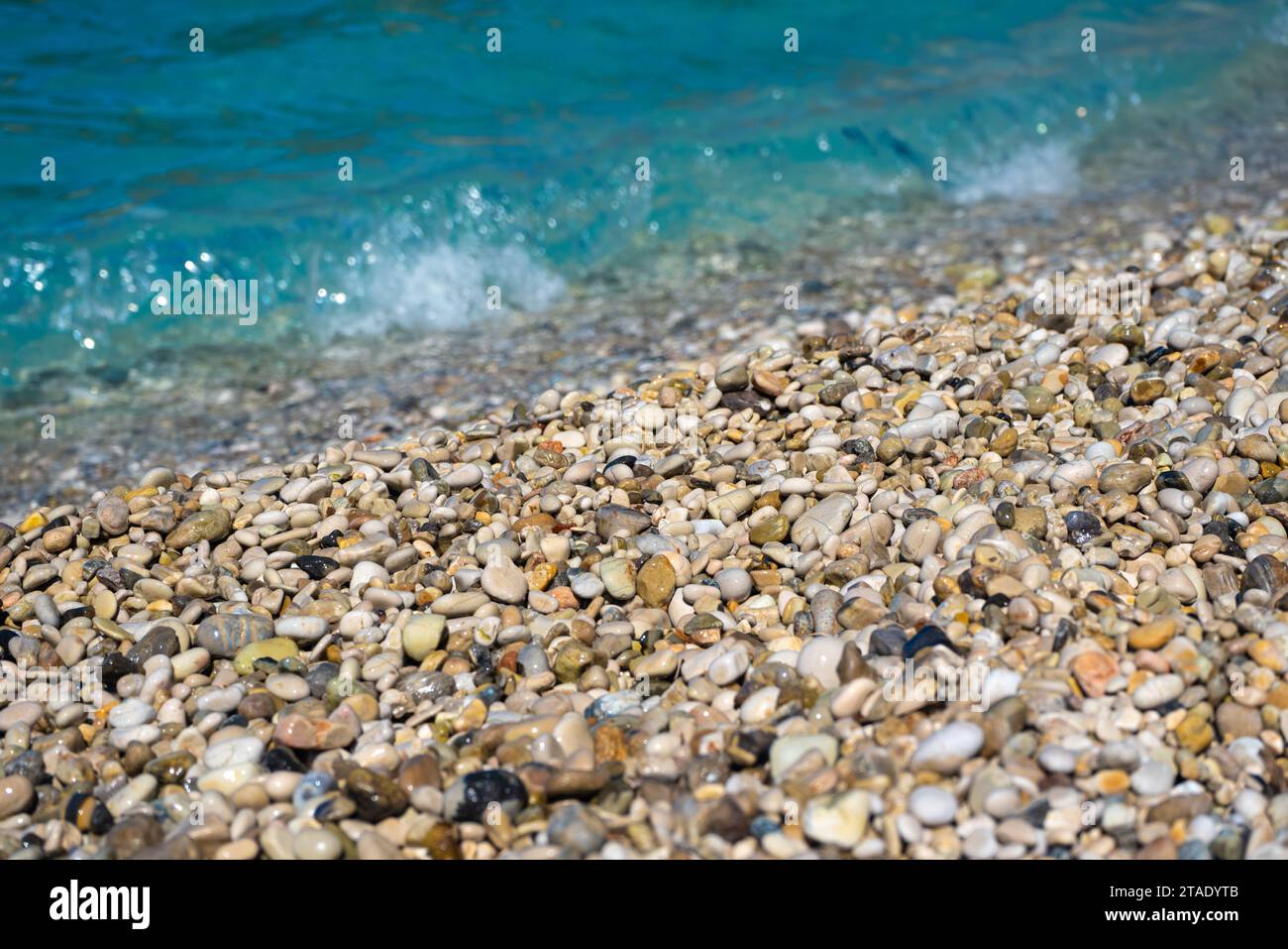 Dettaglio primo piano - piccoli ciottoli o sabbia ruvida su una spiaggia, il sole del pomeriggio sfocato illumina il mare calmo sullo sfondo Foto Stock