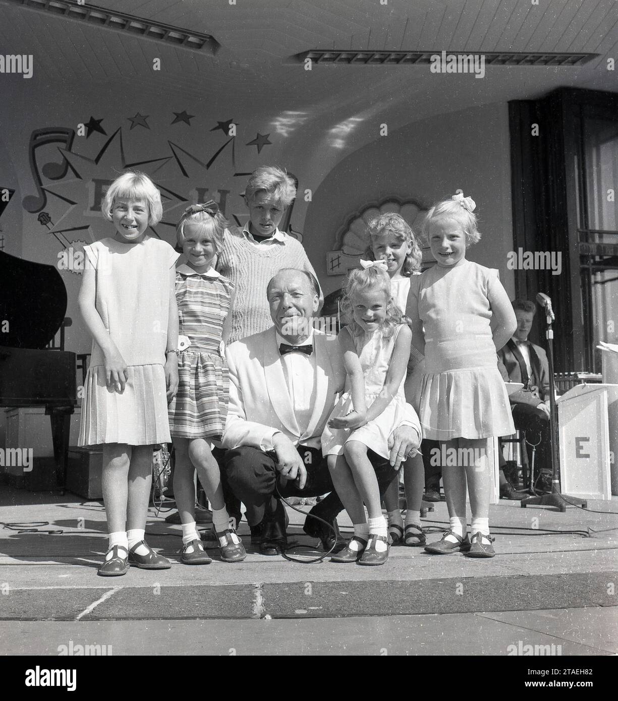 Anni '1960, storico, su un palco un intrattenitore di mare con una giacca bianca e cravatta di fiocco che posa per una fotografia con alcuni bambini piccoli sul palco, Inghilterra, Regno Unito... sorrisi incantevoli sui volti dei giovani. Foto Stock