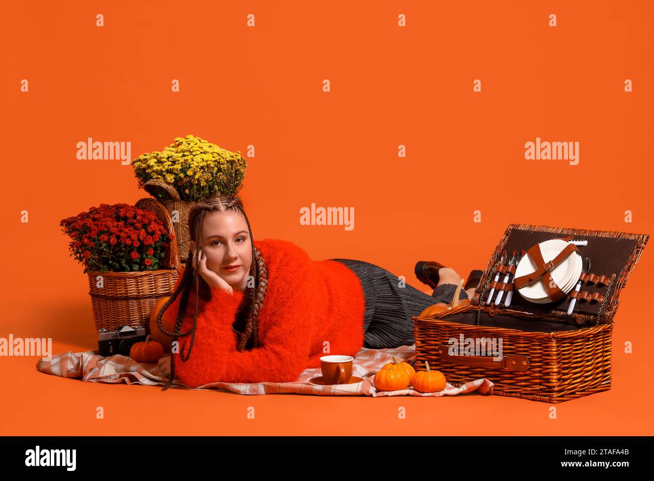 Giovane donna con fiori di crisantemo e zucche che fa picnic su sfondo arancione Foto Stock