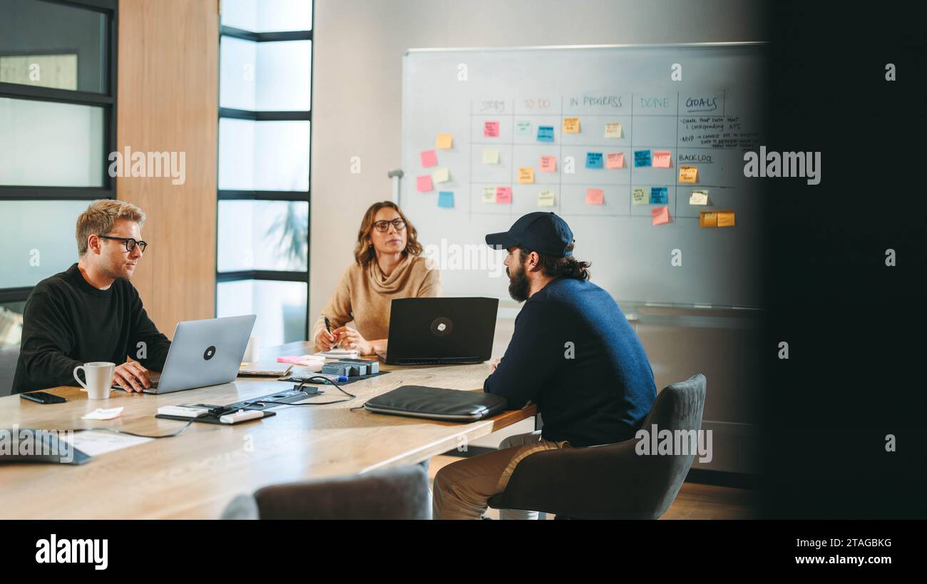 Professionisti aziendali eterogenei in un ufficio moderno si impegnano in una discussione collaborativa intorno a un tavolo da Consiglio di amministrazione. Utilizzo di notebook e puntamento verso una cornice Foto Stock