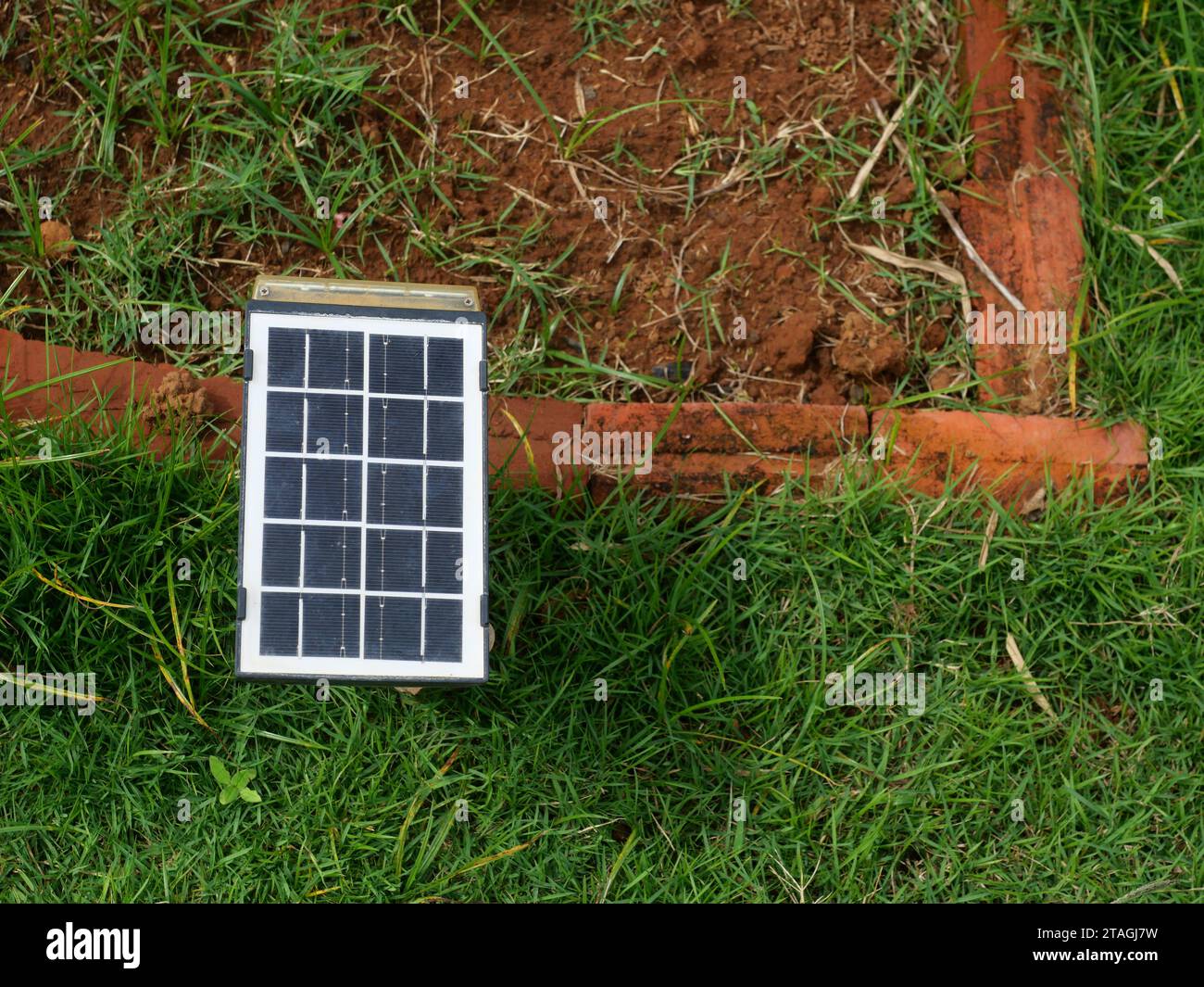 Piccolo pannello a celle solari portatile sul prato con terreno sporco e mattoni rossi sullo sfondo, energia pulita per la generazione di elettricità in casa Foto Stock