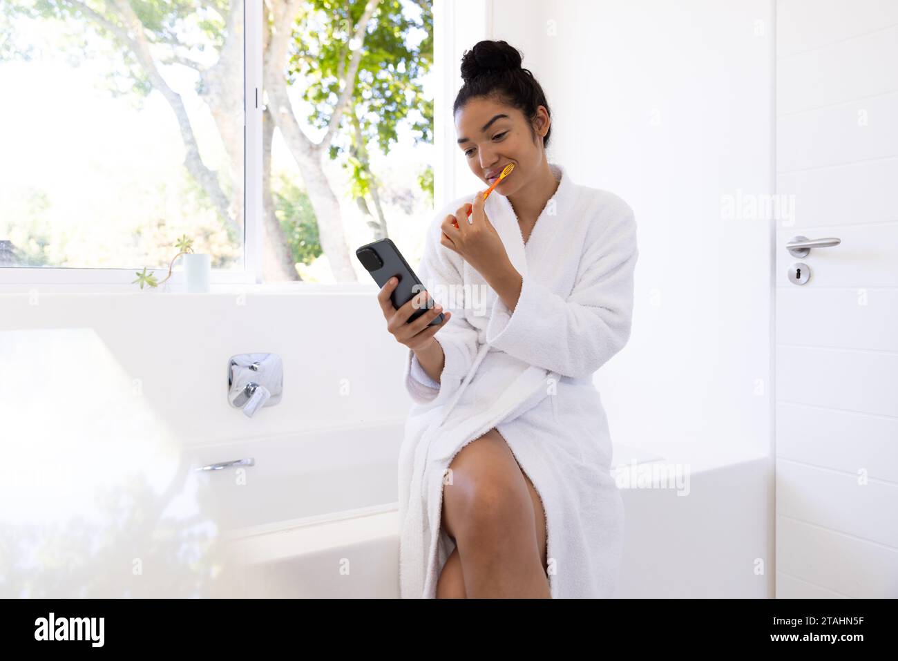 Donna birazziale in accappatoio lavarsi i denti e usare lo smartphone nel bagno soleggiato. Stile di vita, cura di sé, igiene, comunicazione e vita domestica, unale Foto Stock