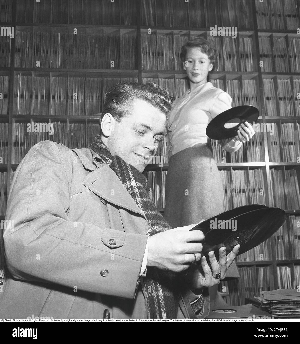 Lars Lönndahl, cantante e attore svedese, è considerato il primo idolo giovanile della Svezia ed è chiamato il più vecchio adolescente della Svezia. Qui con una donna dello staff in un negozio di dischi mentre legge l'etichetta del disco grammofono di Snoddas Nordgren Cupol 4714, in cui vengono registrate le melodie sotto stelle scintillanti e la casa del marinaio. Il posto è un archivio di dischi ben fornito dove gli scaffali sono pieni di dischi grammofonici. Era comune per le persone ascoltare i dischi nel negozio prima di acquistare. 1953. Kristoffersson rif. BM96-5 Foto Stock