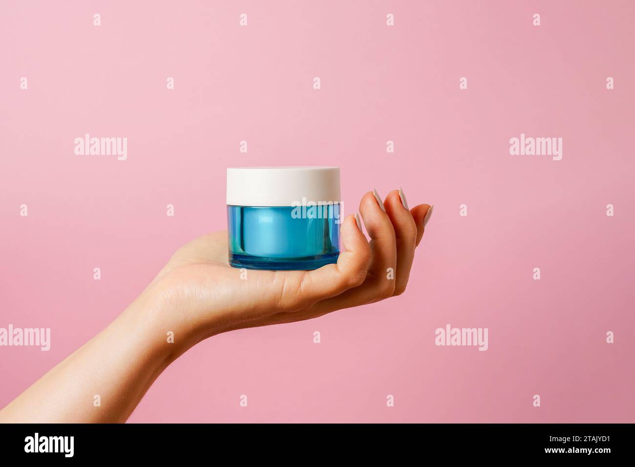 Elegante mano femminile che regge un vaso di vetro blu con crema cosmetica su sfondo rosa. copia spazio Foto Stock