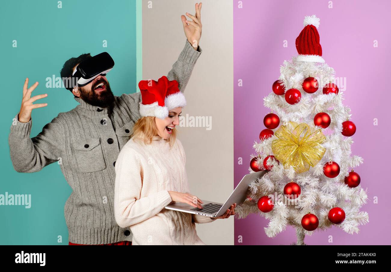 Felice coppia di Natale vicino all'albero di Natale. Donna sorridente nel cappello di Babbo Natale con notebook e felice uomo barbuto con occhiali per realtà virtuale a Natale. 3d. Foto Stock