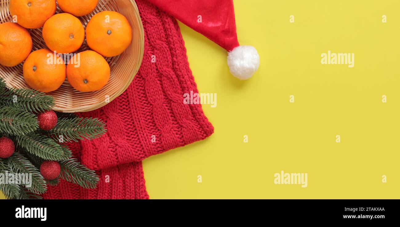 Mandarini in cesto, sciarpa in maglia rossa, cappello di Babbo Natale, rami di abete con palle di Natale su sfondo giallo. Composizione natalizia, anno nuovo, banne Foto Stock