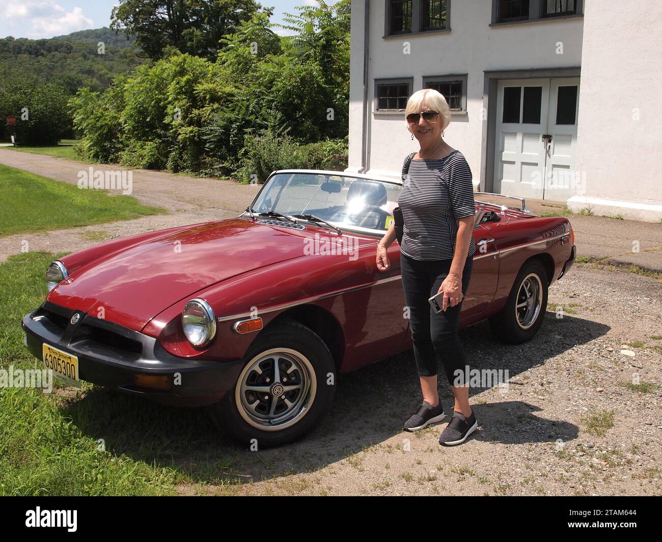 Donna di mezza età in piedi accanto a un roadster da 1977 MGB in Carmine Red. La capote è abbassata per godersi il sole. Foto Stock