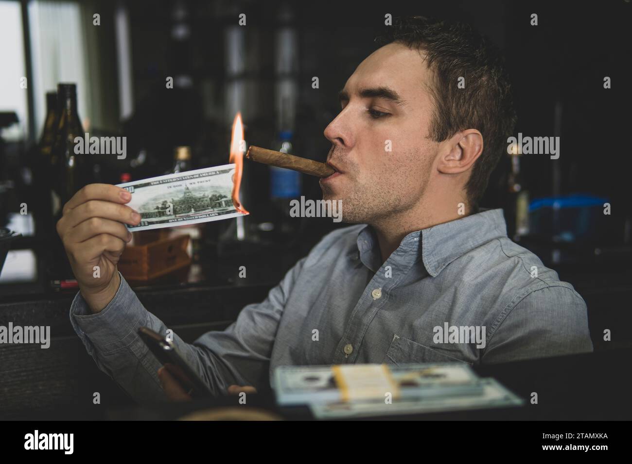 Giovane bell'uomo che accende il suo sigaro con una banconota da 100 dollari sullo sfondo delle bottiglie del bar. mafia russa Foto Stock