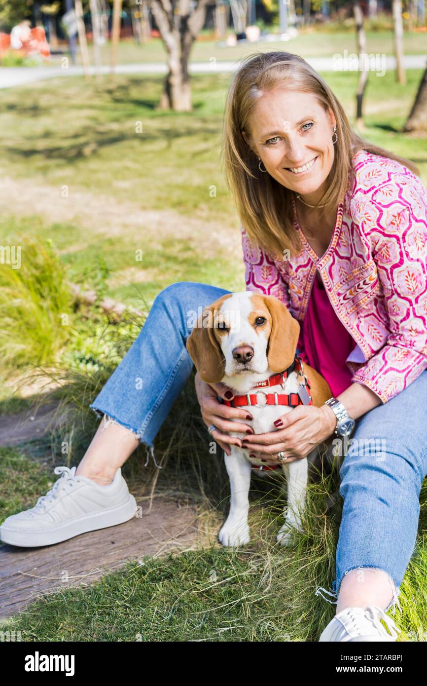 Felice donna adulta bionda sorridente per la macchina fotografica e abbracciando un cane Beagle seduto sull'erba verde al parco Foto Stock