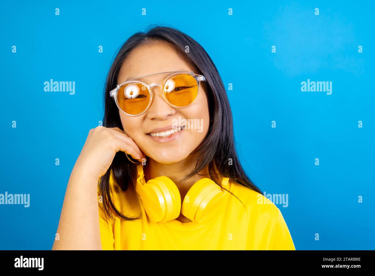 Foto in studio con sfondo blu di una timida e carina donna cinese che indossa occhiali da sole sorridenti alla macchina fotografica Foto Stock