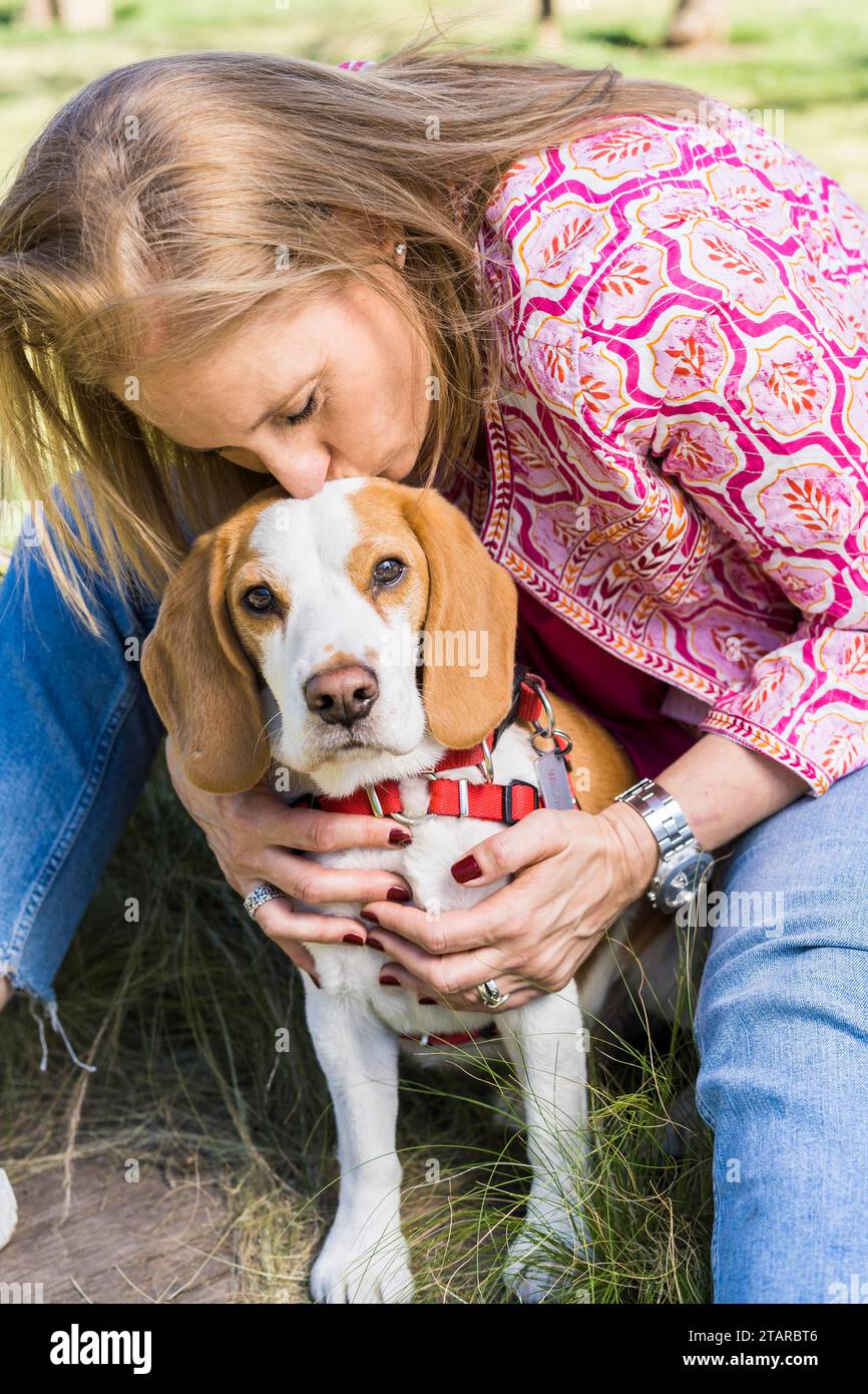 Bionda donna adulta che bacia il suo cane Beagle mentre guarda la macchina fotografica al parco Foto Stock