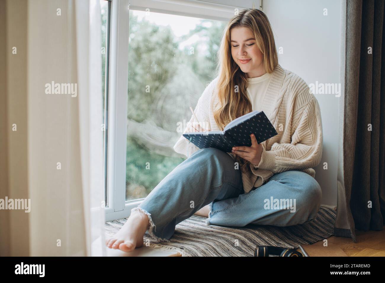 Una giovane donna creativa scrive un piano di memoria e un'idea nel notebook mentre è seduta vicino alla finestra a casa. Concetto di rivista creativa o diario. Foto Stock