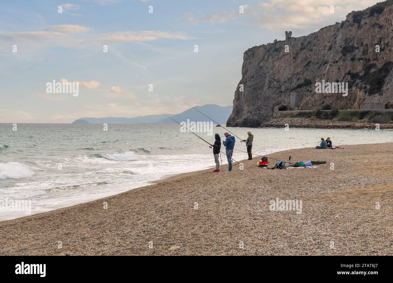 Uomini pesca con canne sulla spiaggia con il promontorio di Caprazoppa e la costa sullo sfondo al tramonto, finale Ligure, Savona, Liguria, Italia Foto Stock