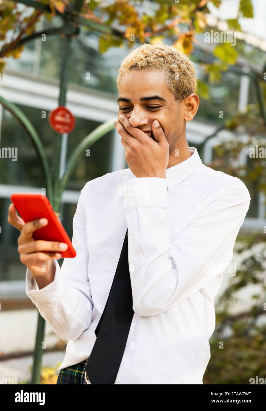 Giovane giovane sorridente con la mano sulla bocca che legge un messaggio del cellulare per strada, foto verticale Foto Stock