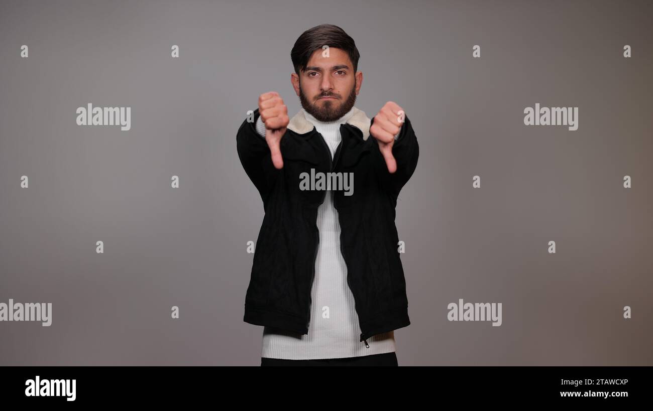 Un giovane che posa con il pollice verso il basso ed esprime antipatia su sfondo grigio. Foto Stock