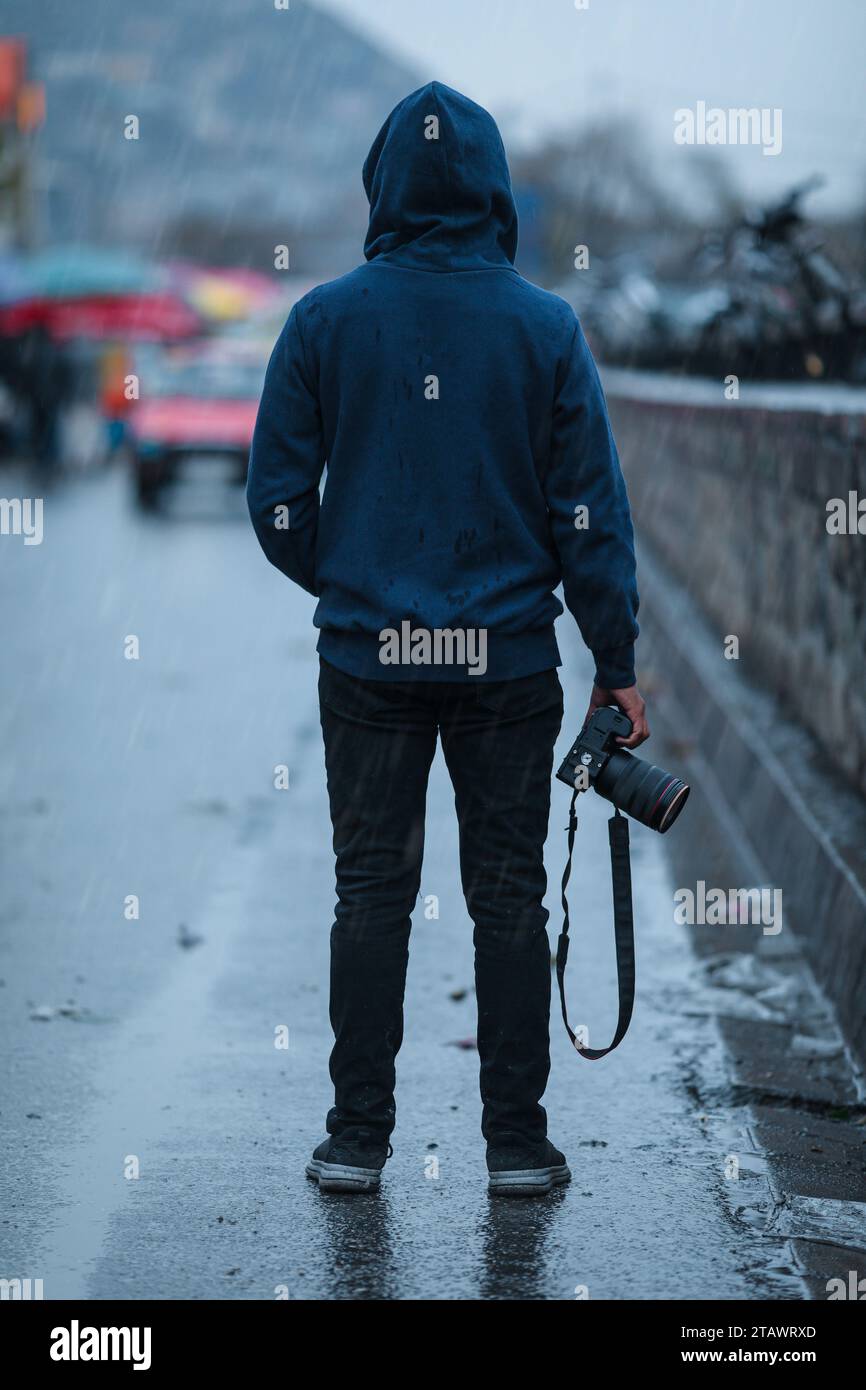 Un fotografo che indossa una felpa con cappuccio, regge una macchina fotografica e si trova in strada. Foto Stock