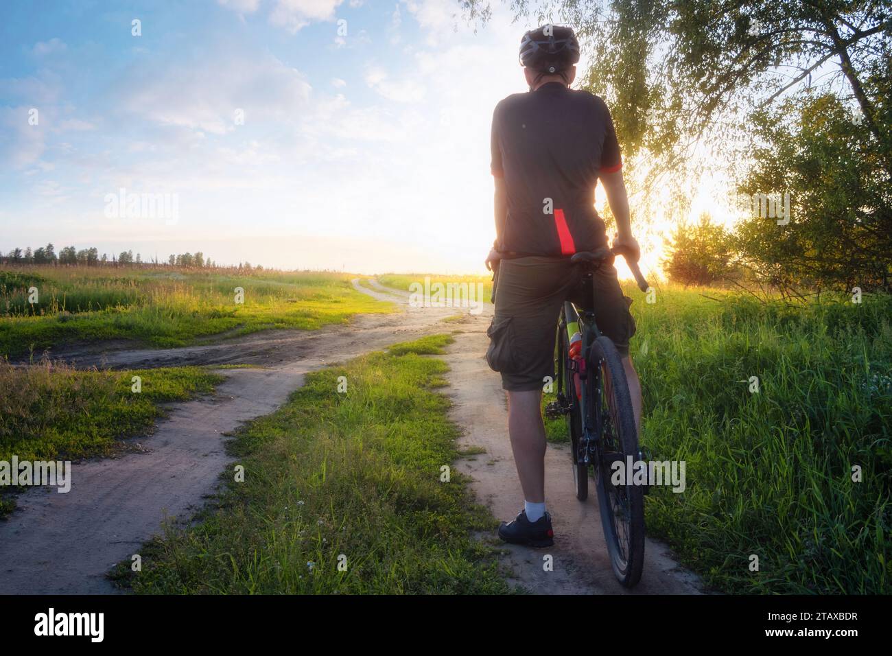 Uomo che cavalca una bicicletta di ghiaia sul sentiero al tramonto. Paesaggio colorato con ciclista, bicicletta, campo, erba verde, alberi e luce solare. Sport e tr Foto Stock
