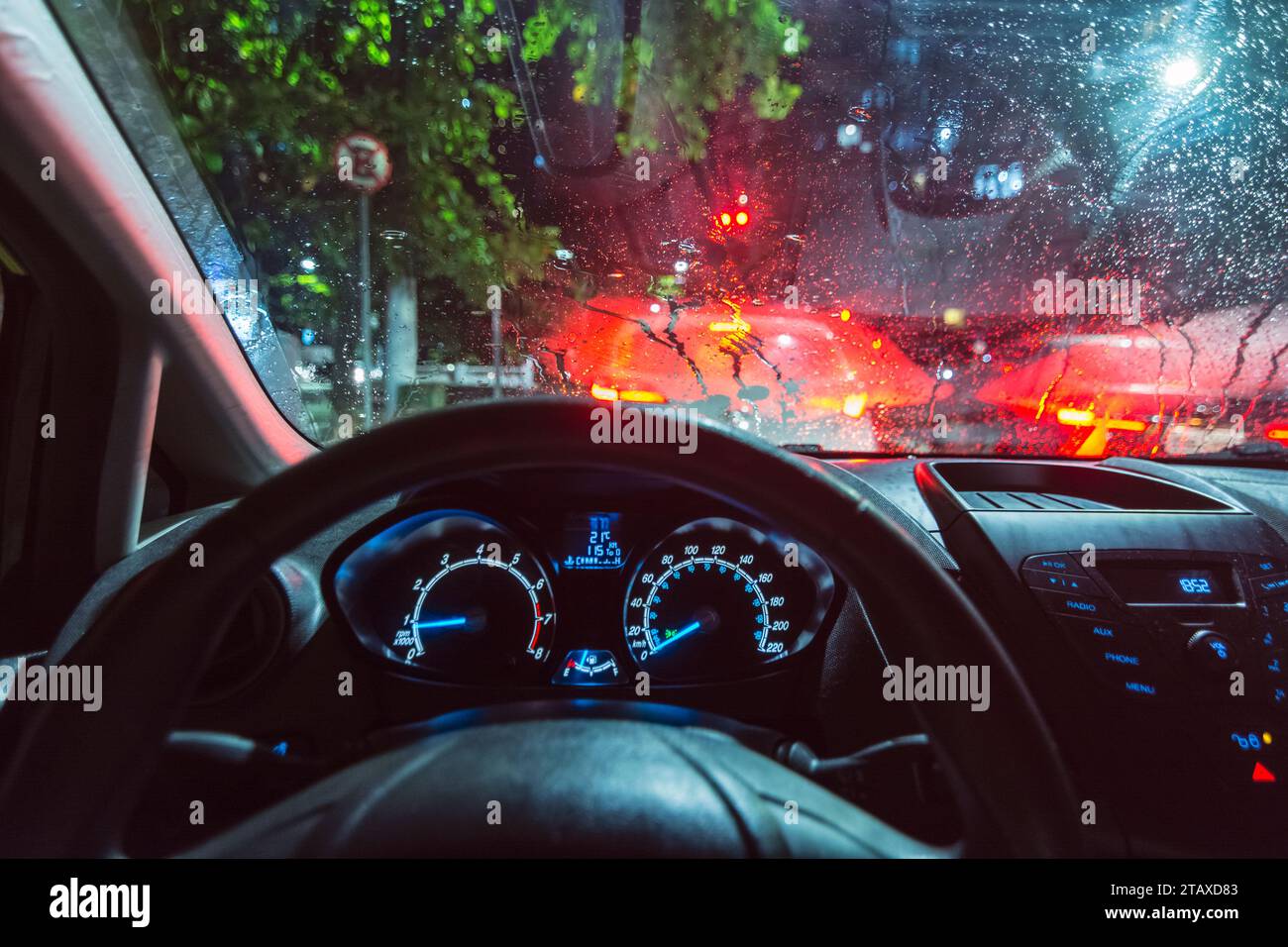 Vista interna di un'auto, dal punto di vista del conducente, si è fermata al semaforo in una giornata di pioggia. Luci plancia, parabrezza a goccia di pioggia. Foto Stock