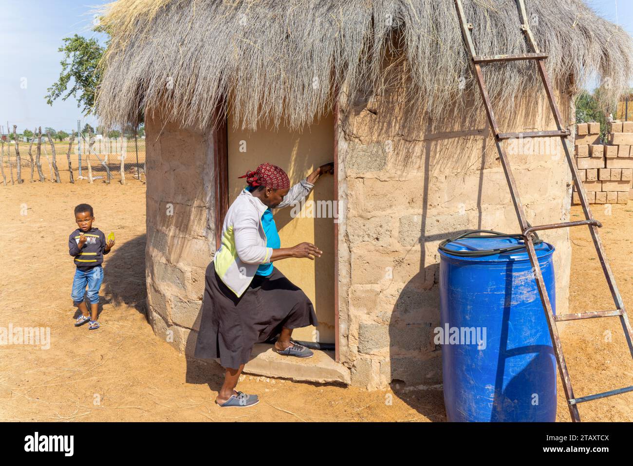 villaggio africano, vecchia donna che apre la porta alla sua capanna rondavel, ragazzino che mangia uno spuntino guardando sua nonna Foto Stock