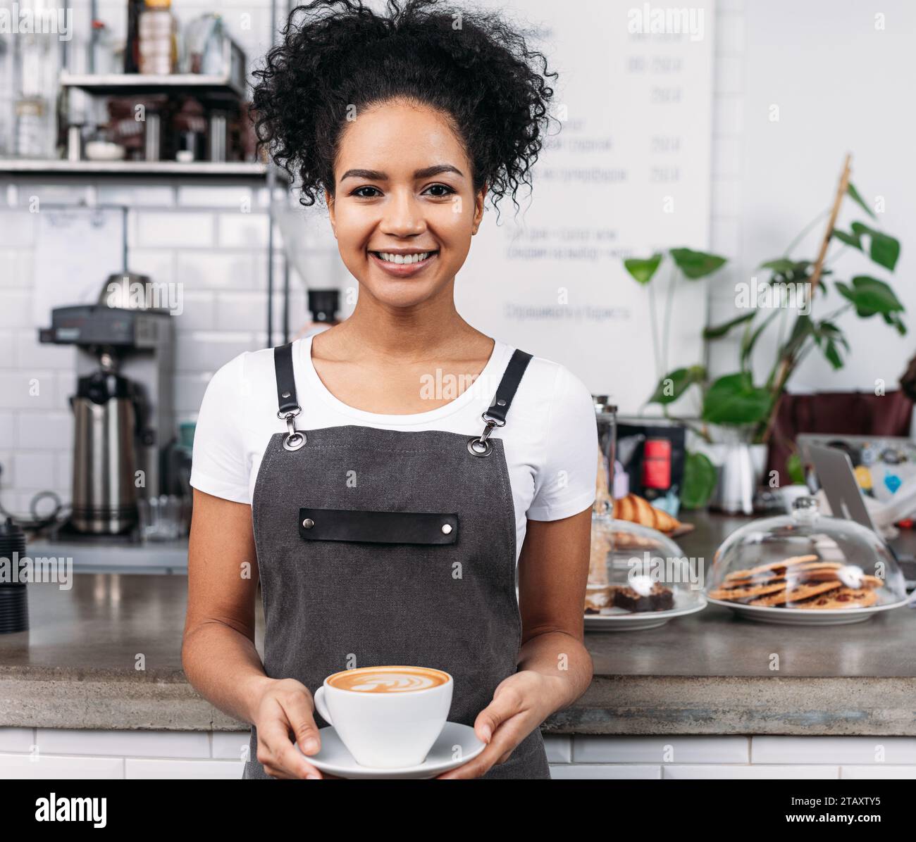 Cameriera sorridente con una tazza di caffè. Donna afro-americana che lavora come barista e tiene in mano una tazza con un cappuccino appena preparato. Foto Stock