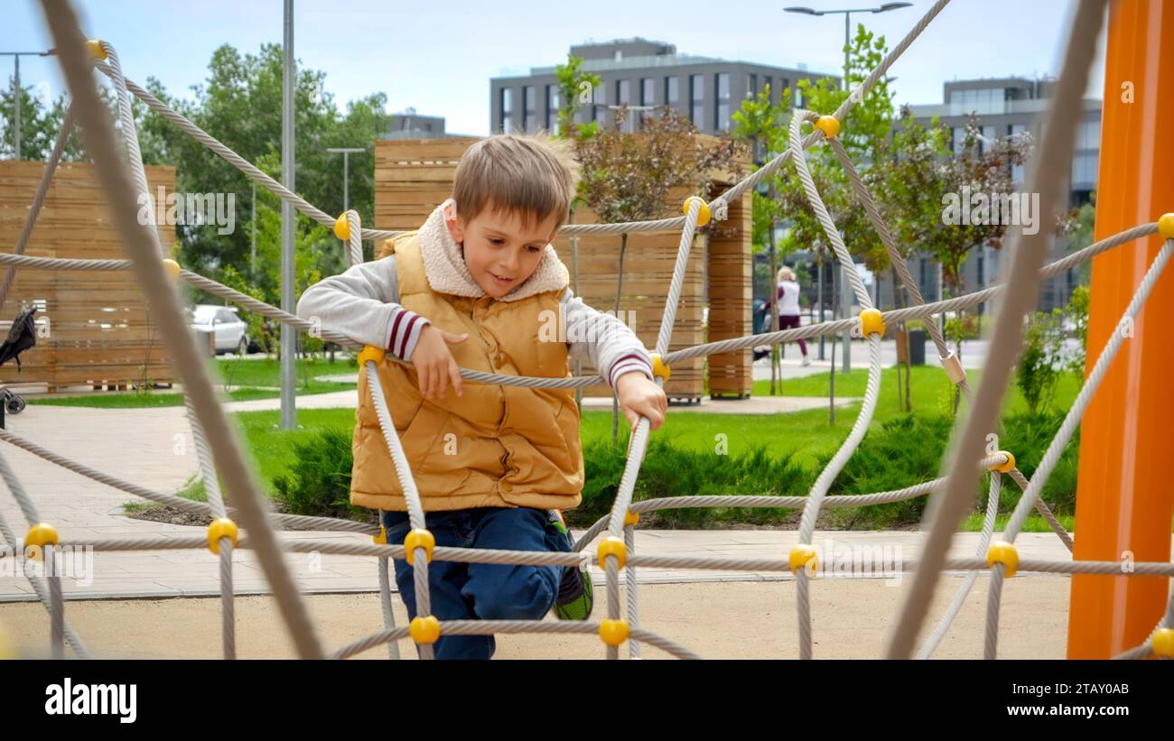 Un ragazzo allegro che si arrampica tra corde e ostacoli sul parco giochi all'aperto in un moderno edificio. Foto Stock