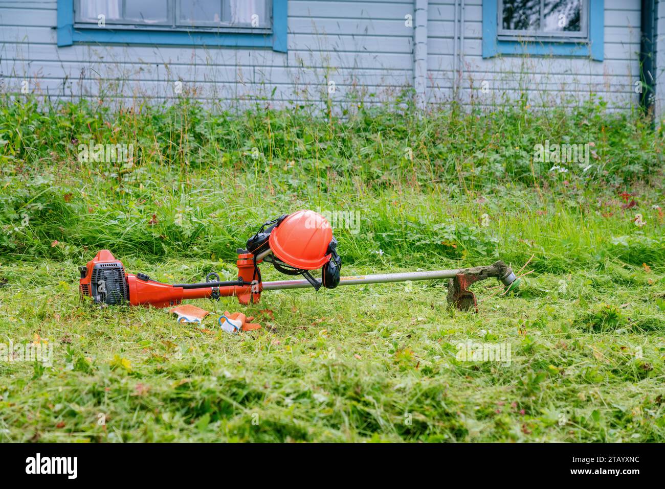 Rifinitore arancione per corde a gas, casco di sicurezza con protezione per gli occhi e guanti gommosi su erba fresca di fronte a cottage di montagna in legno. Ravvicinato via Foto Stock
