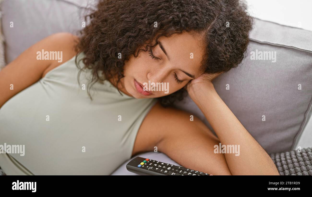 La giovane donna ispanica esausta trova comfort sul divano, si addormenta nell'accogliente sottofondo del suo salotto a casa, lasciando il telecomando Foto Stock