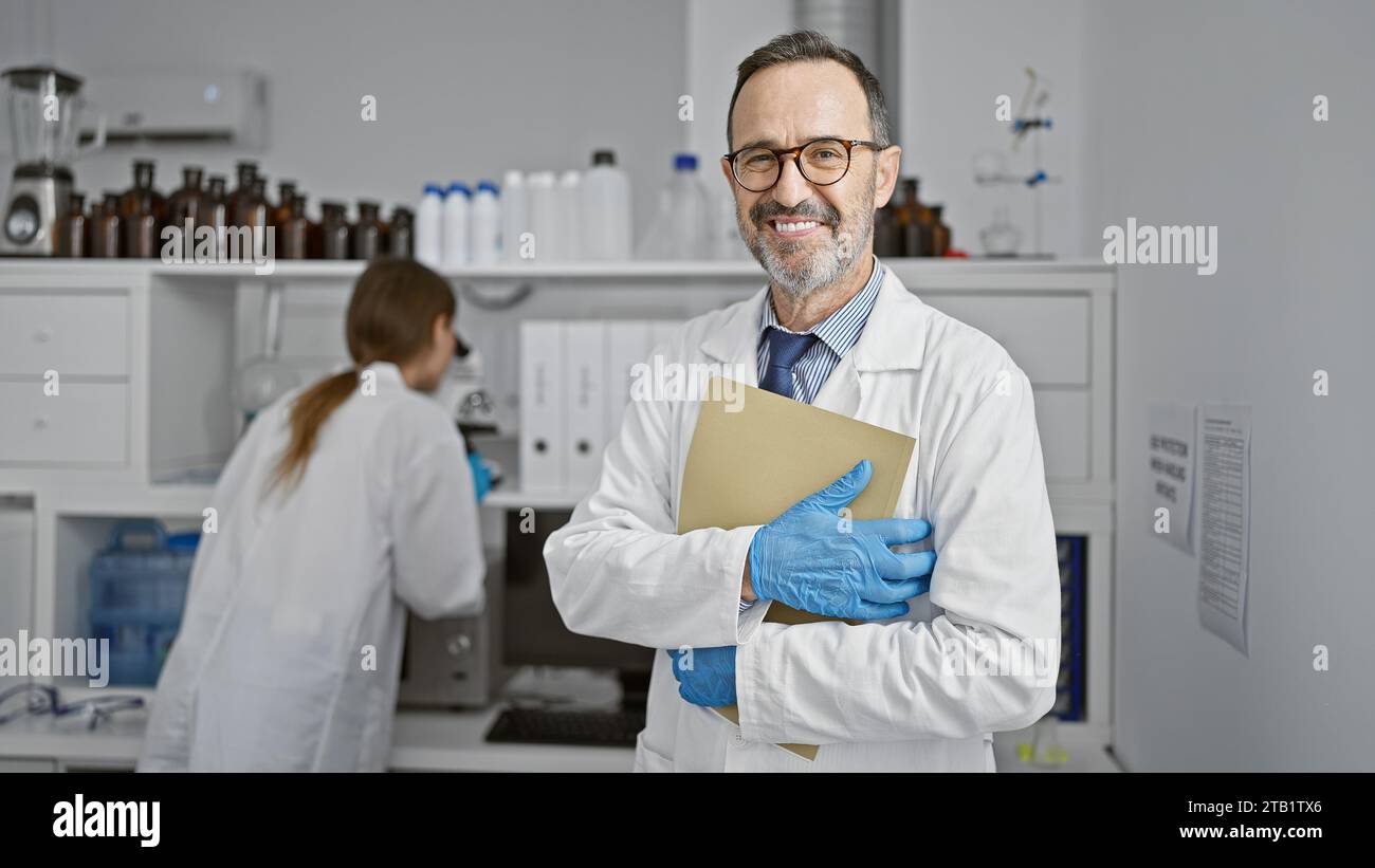 Due scienziati sorridenti, partner del crimine, in piedi con le braccia incrociate in laboratorio, a destreggiarsi con i documenti del loro ultimo sbalorditivo Foto Stock