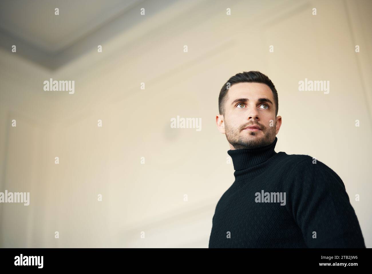 Uomo pensivo con dolcevita nera che guarda verso l'alto in una stanza spaziosa e luminosa. Ritratto di bellezza maschile Foto Stock