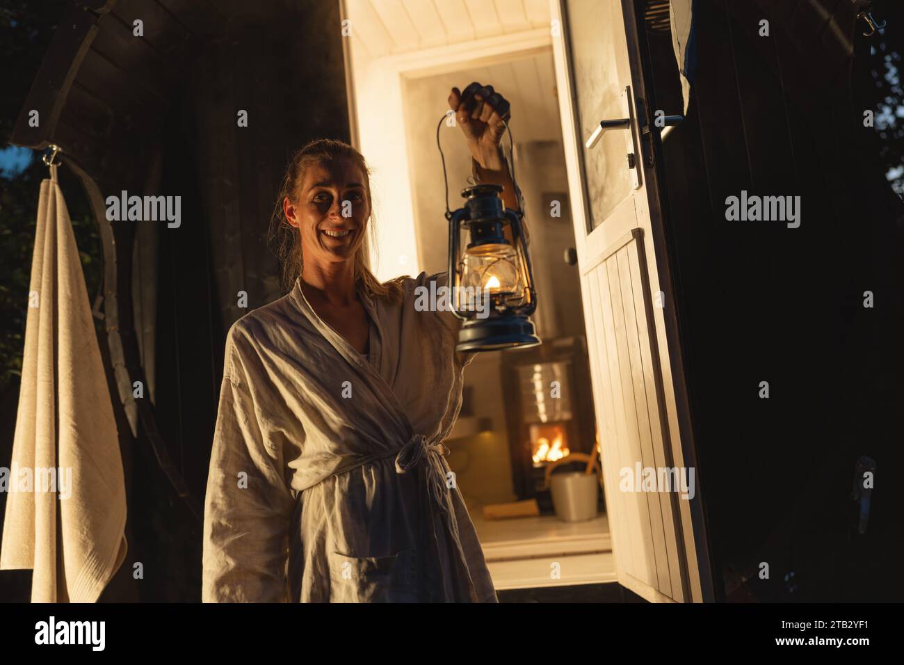 Donna sorridente che tiene una lanterna all'ingresso di una sauna finlandese a botte durante la serata. Foto Stock