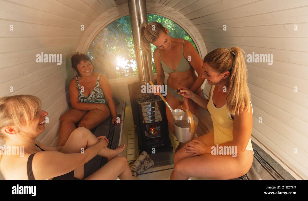 Quattro donne in una sauna, una che gocciola il profumo della sauna in un mestolo d'acqua sopra il riscaldatore. Immagine concetto sauna finlandese mobile Foto Stock