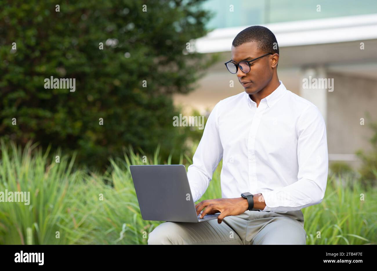 Uomo d'affari nero professionista con occhiali che utilizza un computer portatile mentre è seduto all'esterno Foto Stock