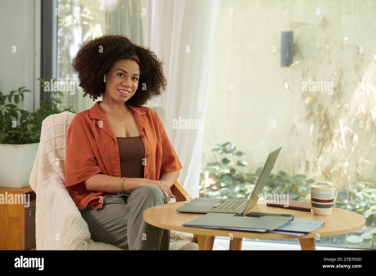 Ritratto di una donna sorridente seduta su un tavolino da caffè con un computer portatile aperto, che lavora da casa Foto Stock