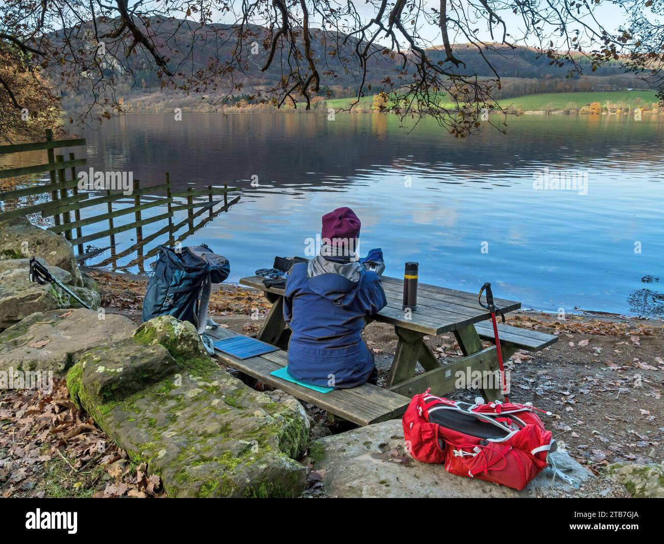 Donna seduta al tavolo da picnic in legno sulle rive del lago Ullswater nell'English Lake District, vista da Sandwick in autunno, Cumbria, Inghilterra, Regno Unito Foto Stock