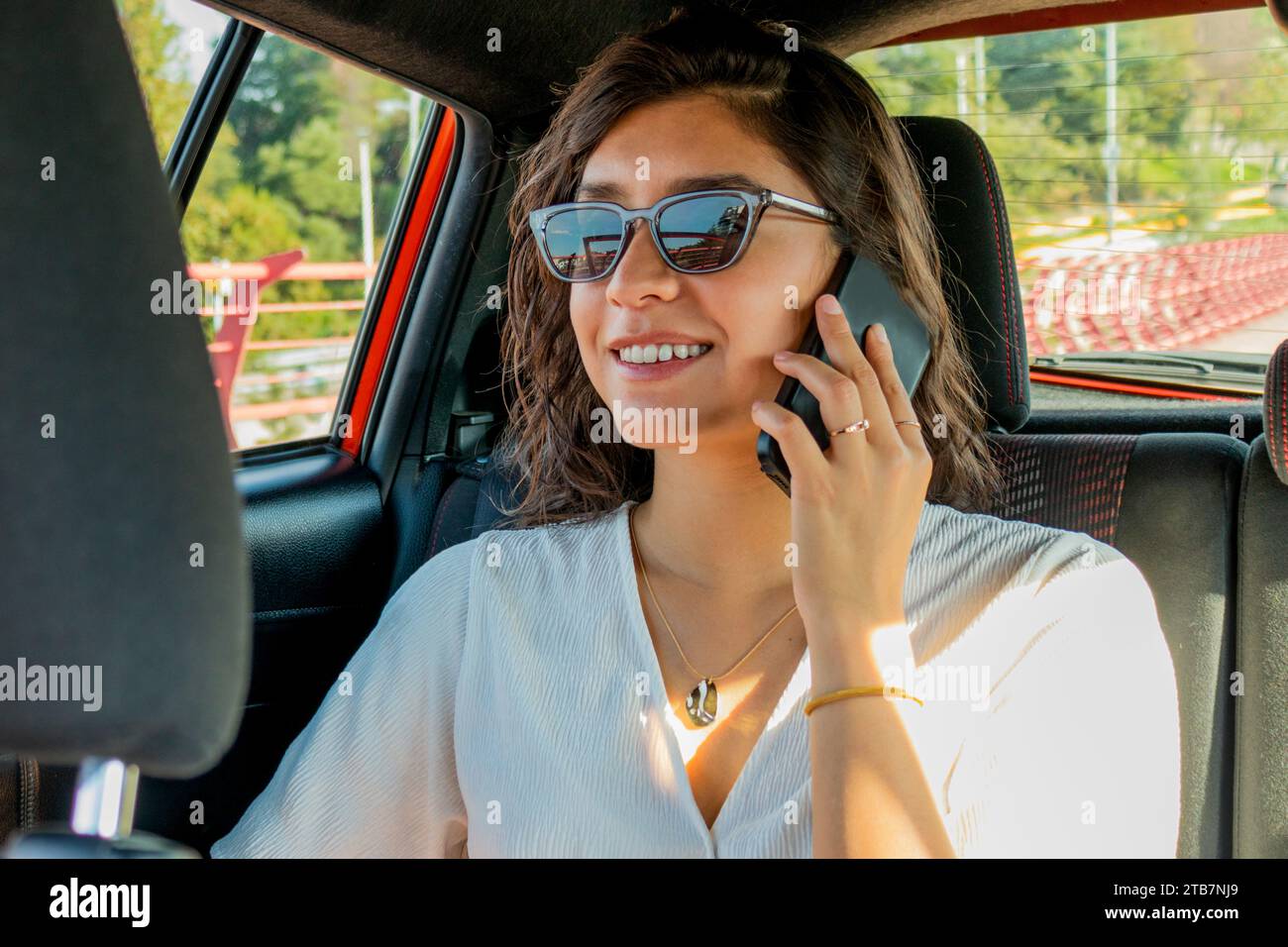 Sorridente bella giovane donna caucasica che indossa occhiali da sole e casuali che ascolta lo smartphone durante una chiamata mentre si siede sul sedile posteriore dell'auto Foto Stock