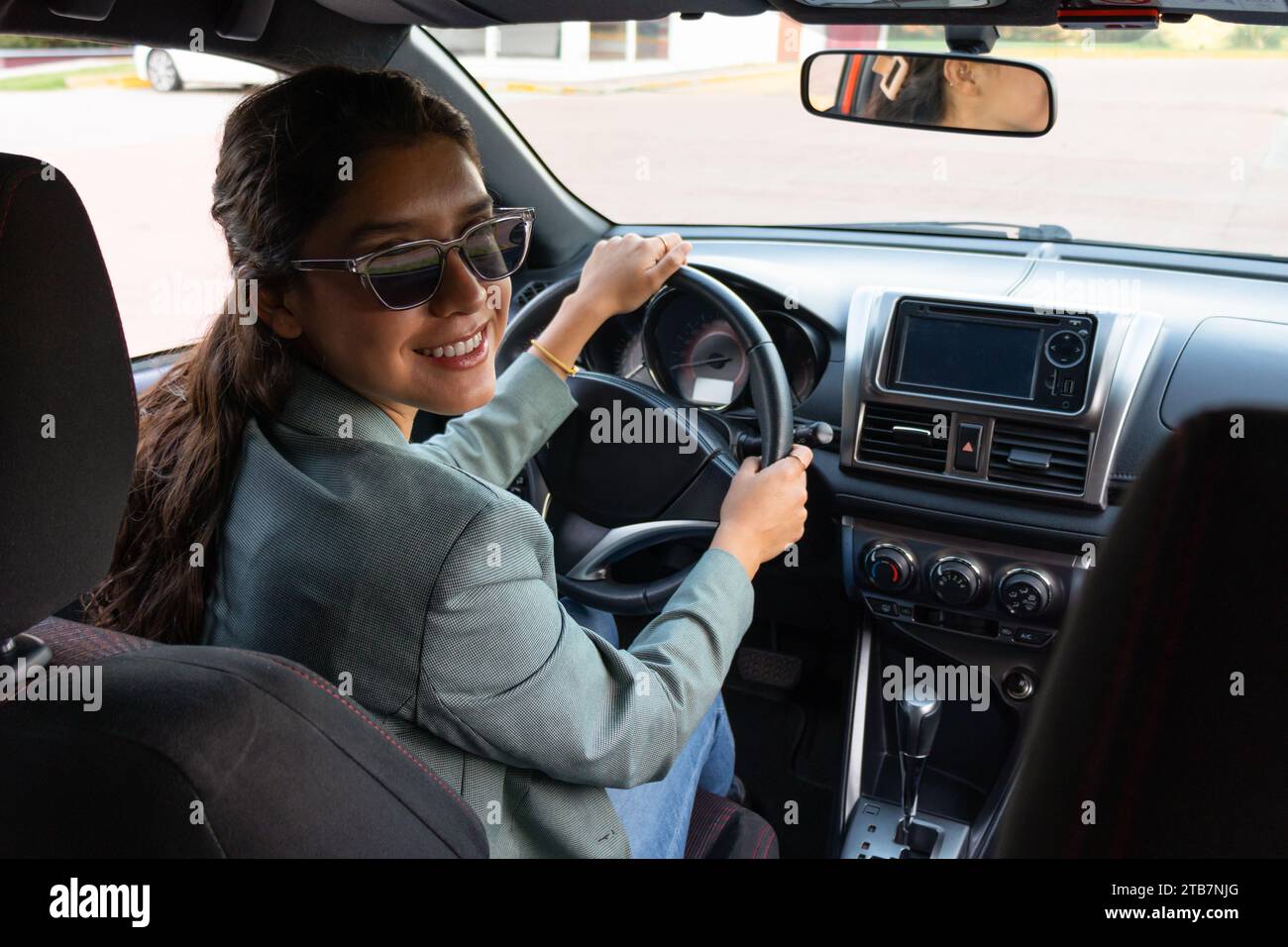 Ritratto di un giovane e allegro dirigente caucasico che indossa occhiali da sole e blazer che guarda a spalla mentre guida un'auto moderna in città Foto Stock