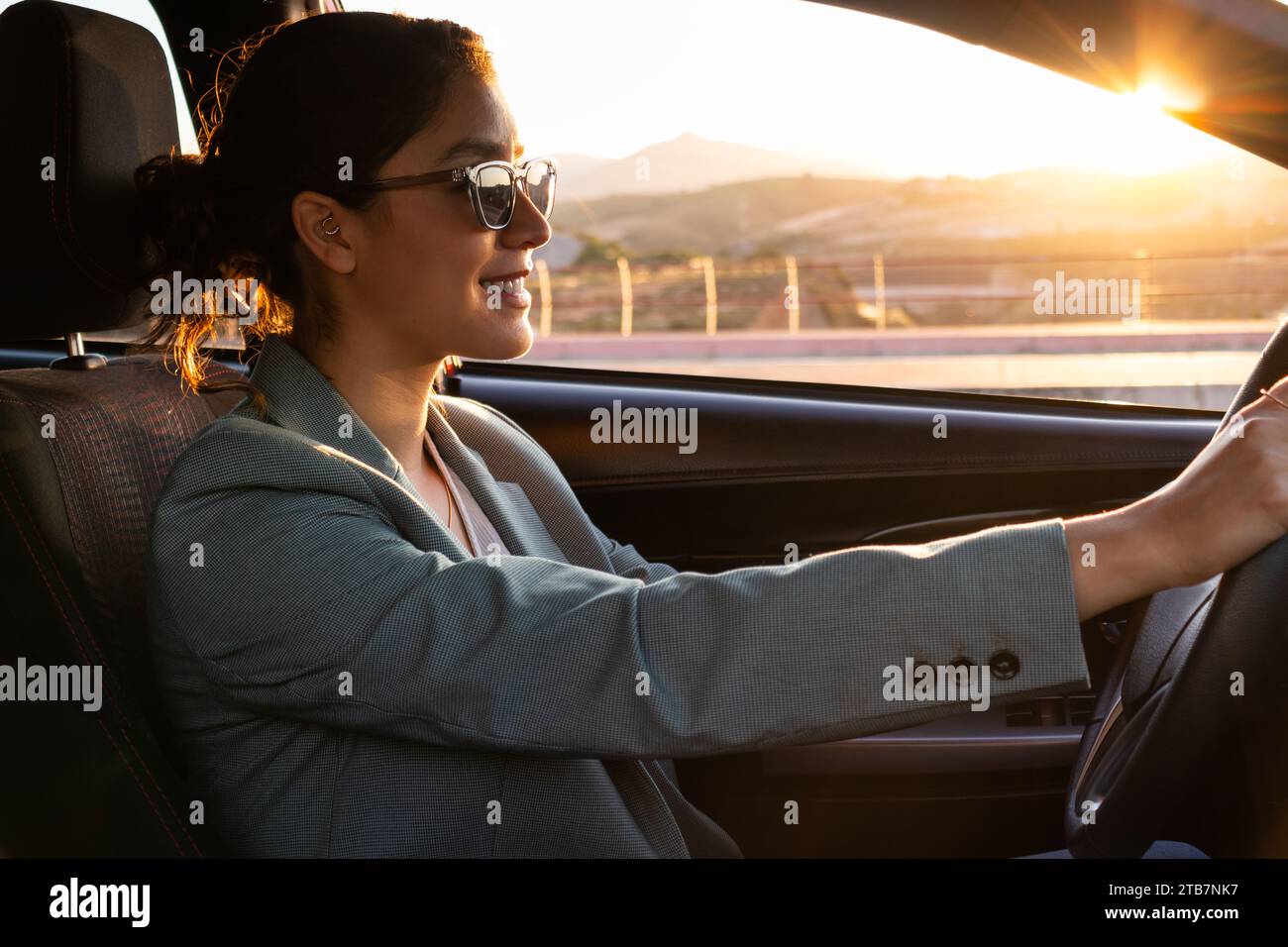 Ritratto di un giovane e allegro dirigente caucasico che indossa occhiali da sole e blazer che guida un'auto moderna al tramonto in città Foto Stock