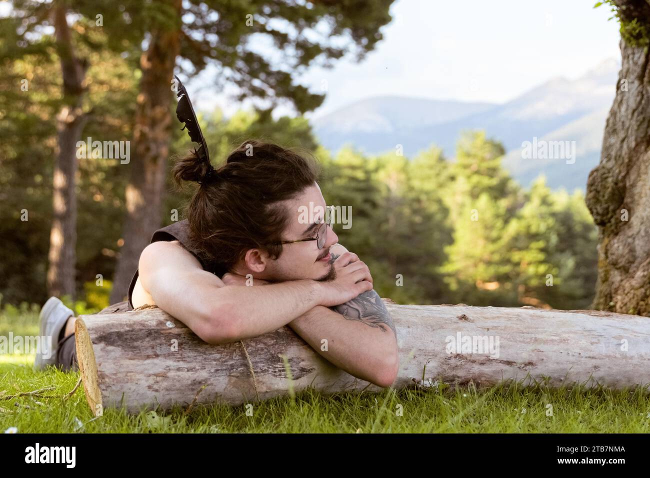 Un uomo si stende su un tronco di albero caduto in una radura forestale osservando montagne lontane Foto Stock
