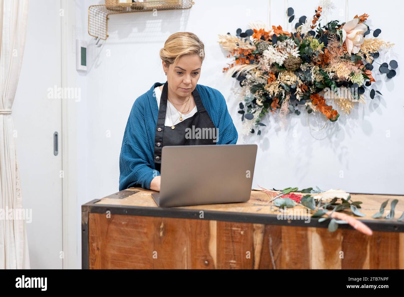 Donna d'affari concentrata che lavora su un computer portatile a una scrivania rustica in legno con una disposizione floreale decorativa sullo sfondo Foto Stock