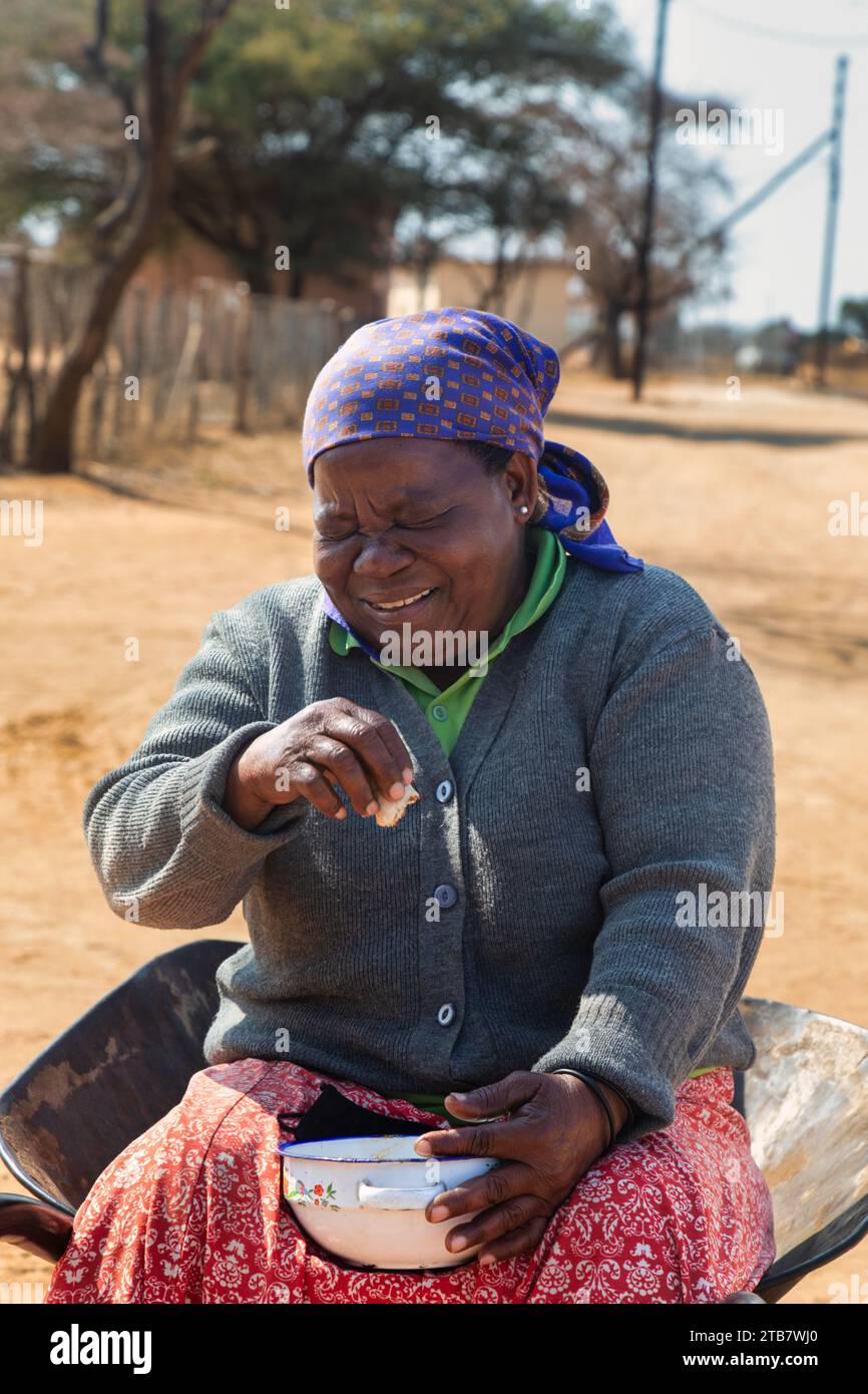 villager, una vecchia donna africana in una carriola che mangia del cibo tradizionale e ride di una buona battuta Foto Stock