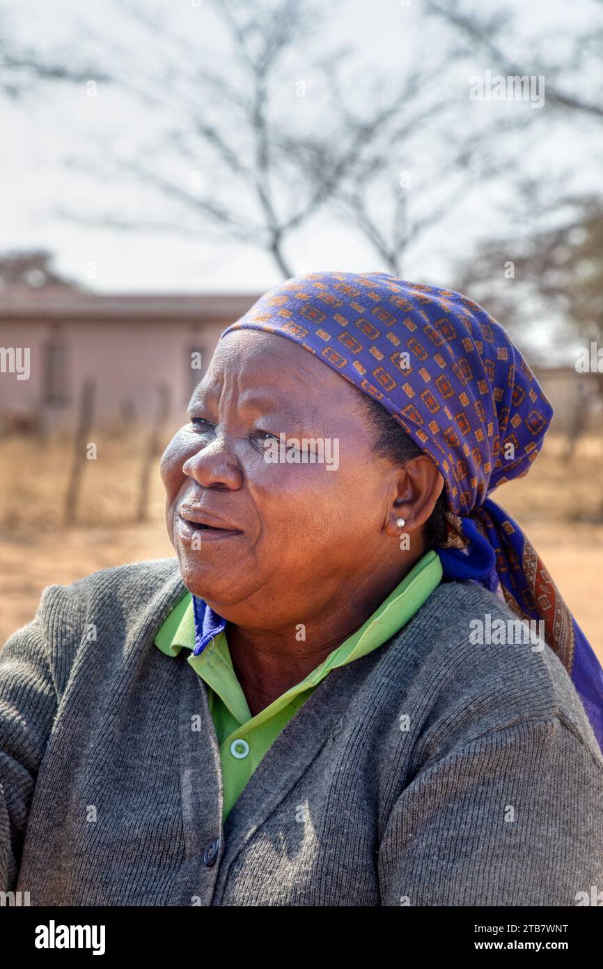 villager , colpo di testa di vecchia donna africana con sciarpa blu, casa e strada sterrata sullo sfondo Foto Stock