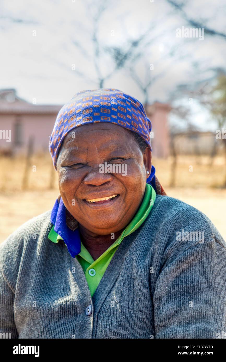 villager, vecchia donna africana che rideva di una bella battuta, di una casa e di una strada sterrata sullo sfondo Foto Stock