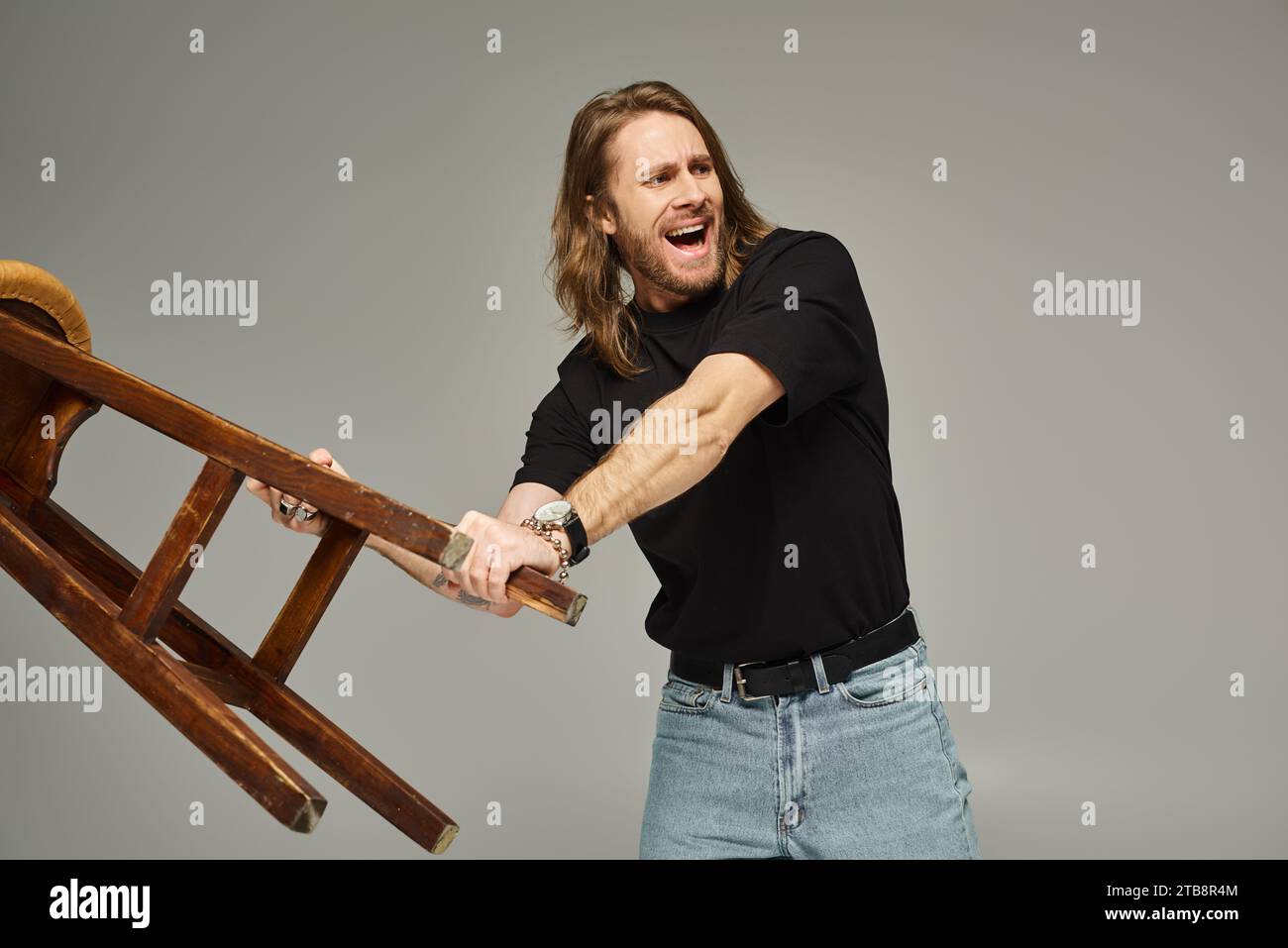 uomo emotivo con capelli lunghi in jeans e t-shirt che porta sgabello alto e urla grigia Foto Stock